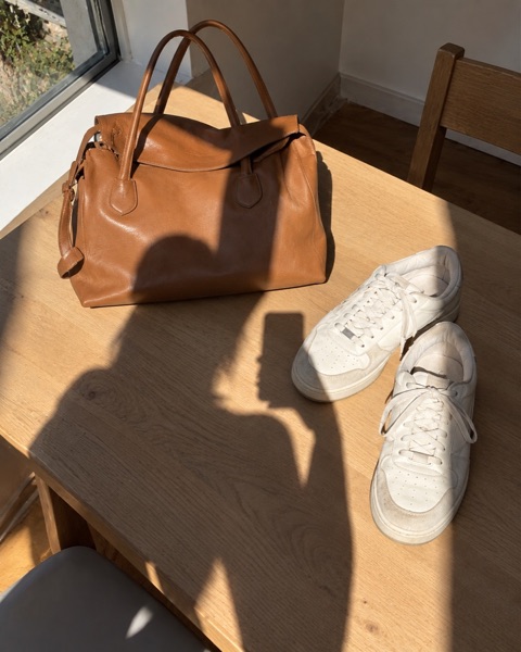 A leather bag and white sneakers on a table with the photographer's shadow across them